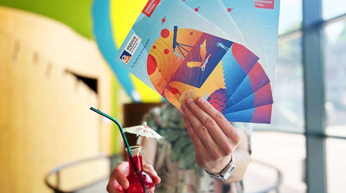 personne tendant devant lui les flyer de l'été et un ballon rempli d'eau coloré en rouge